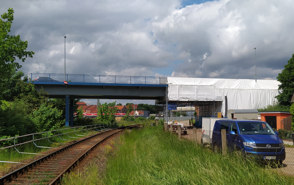 Eine blaue Stahlbrücke, auf der eine Auot fährt, führt über Eisenbahnschienen. Auf der rechten Seite ist ein Teil der Brücke mit einem weißen Baustellenzelt eingehaust. Unter der Brücke steht rechts neben den Bahnschienen ein Zaun, an dem ein Schotterweg entlang führt. Auf dem Weg steht ein blauer Transporter mit Anhänger.