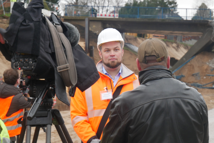 Ein Mann mit orangener Jacke und Bauhelm steht vor einer Kamera und gibt ein Interview. Im Hintergrund ist eine Baustelle.