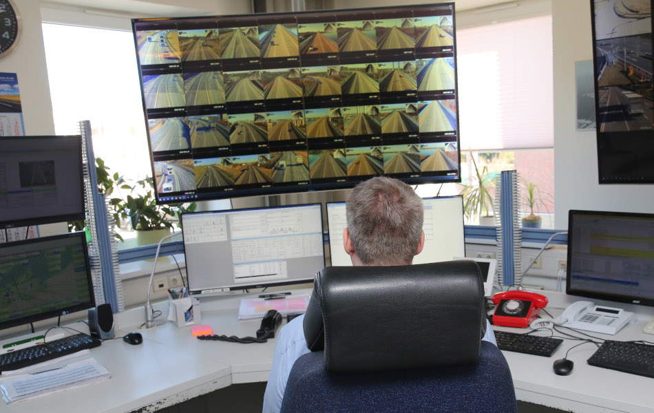 Ein Mann sitzt auf einem Bürostuhl und blickt auf mehrere Monitore, darunter ein ganz großer Bildschirm mit vielen verschiedenen Kameraperspektiven.