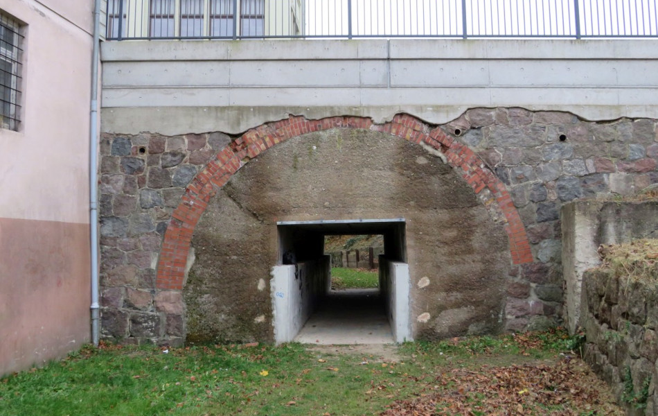 Unter einer Brücke aus verschiedenen Gesteinsarten und Mauerwerken führt ein Tunnel für Fußgänger*innen hindurch.