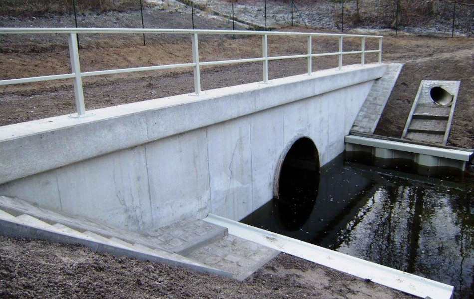 Eine Betonbrücke mit grauem Geländer führt über ein Gewässer. Am Rand der Brücke befinden sich Stufen und Flächen mit Pflastersteinen.