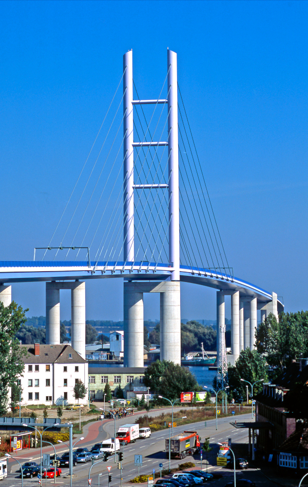 Hinter einer Straße und Wohnhäusern ragen große Brückensäulen aus einem Gewässer. Oben auf der Brücke steht ein riesiger Pylon, von dem Metallstreben zu den Brückenrändern verlaufen.