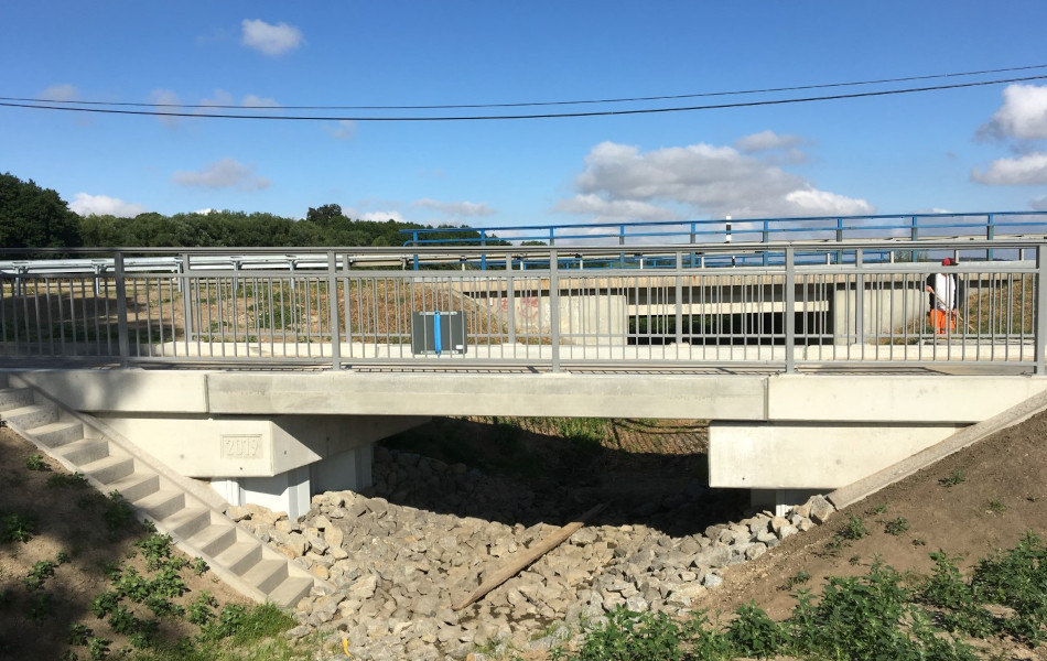 Eine Brücke, die aus mehreren Betonteilen besteht und ein graues Geländer hat, führt über einen Graben. In dem Graben liegen zahlreiche Steine. Im Hintergrund ist eine größere Straßenbrücke.