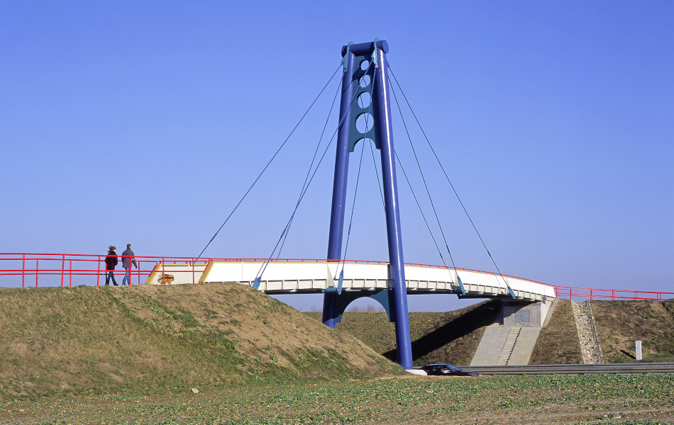 Eine Frau und ein Mann mit einem Hund gehen über eine Brücke, die über einen blauen Pylonen verfügt. Die Brückengeländer sind rot.