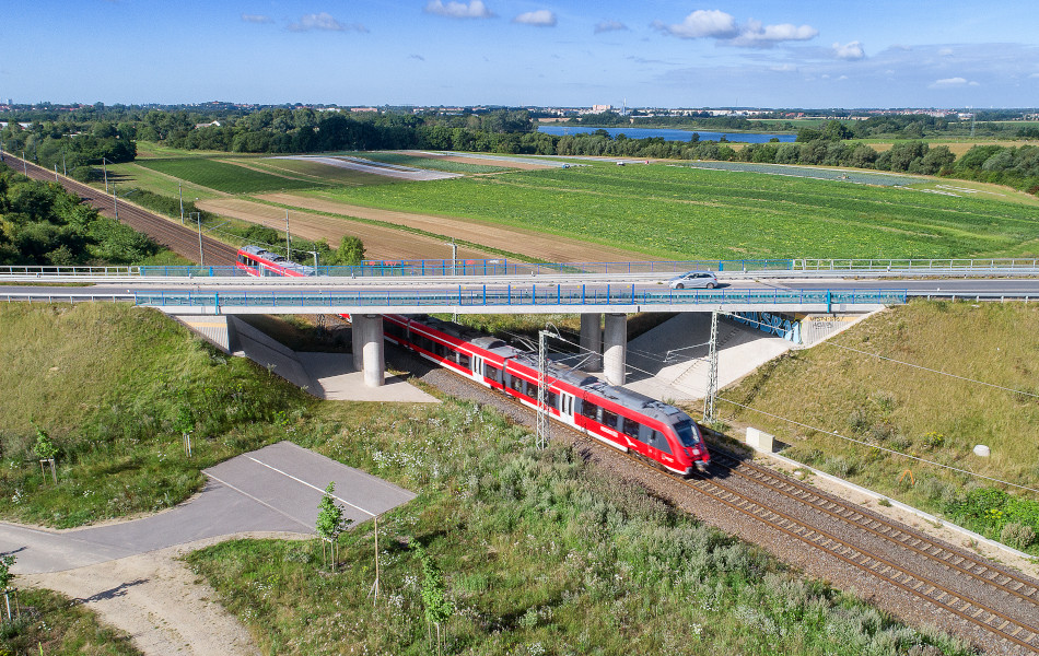 Ein Auto fährt auf einer Brücke mit blauen Geländern und Betonpfeilern. Unter der Brücke fährt ein roter Zug auf Bahnschienen hindurch.