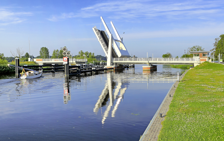 Eine flache Brücke führt über einen Fluss und ist in der Mitte aufgeklappt. An einem Steg liegt ein Boot.