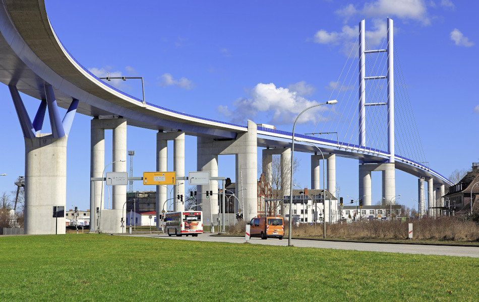 Auf einer Straße fährt ein Bus und ein Transporter. Darüber erhebt sich auf Betonpfeilern eine riesige Brücke.