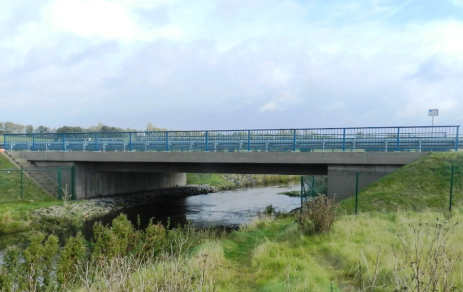 Unter einer Straßenbrücke aus Beton fließt ein Fluss hindurch. Die Ufer sind begrünt. Das Geländer der Brücke besteht aus blauen Stahl, am Fahrbahnrand stehen auf der Brücke Schutzplanken.