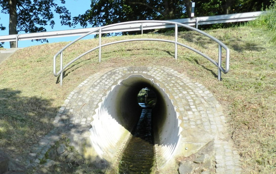 Ein Bach fließt unterhalb einer Straße durch eine kleine Brücke, die mit Pflastersteinen rundlich gemauert ist. Links neben der Brücke führen Treppenstufen zur Straße.