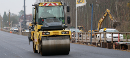 Eine Walze fährt auf einer Baustelle über eine frisch asphaltierte Straße. Im Hintergrund und rechts neben der Straße fahren hinter einer Holz-Absperrung Autos vorbei. Daneben arbeitet ein Bagger. 