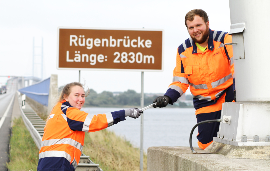 Eine junge Straßenwärterin in orangener Arbeitskleidung reicht einem Kollegen einen Schraubenschlüssel. Im Hintergrund ist die Rügenbrücke. 