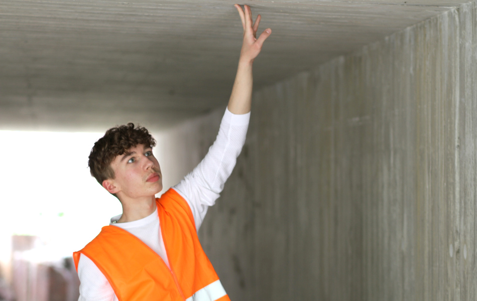 Ein junger Mann mit orangener West blickt nach oben und berührt mit seiner ausgestreckten Hand eine Betondecke.