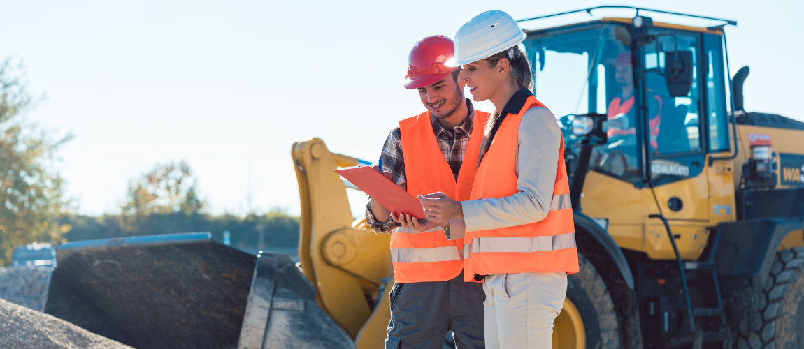 Ein Mann und eine Frau stehen auf einer Baustelle und schauen auf einen Plan. Sie tragen orangene Westen und Bauhelme. Im Hintergrund ist ein Radlader.