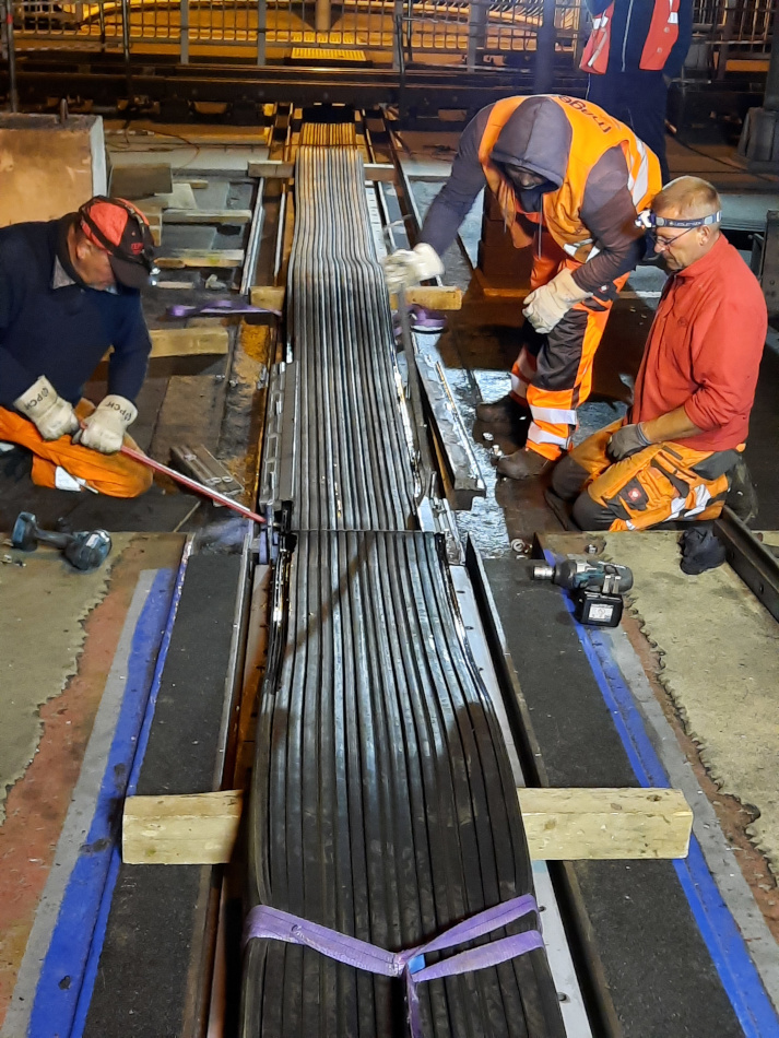 Zwei Bauarbeiter sitzen sich knieend gegenüber. Zwischen ihnen verläuft ein breites Gummiband. Einer der Bauarbeiter drückt das Gummi mit einer Eisenstange in den Übergang zwischen zwei Fahrbahnen. Ein weitere Bauarbeiter, der ihm gegenüber steht, drückt ebenfalls mit einer Eisenstange auf das Gummi.