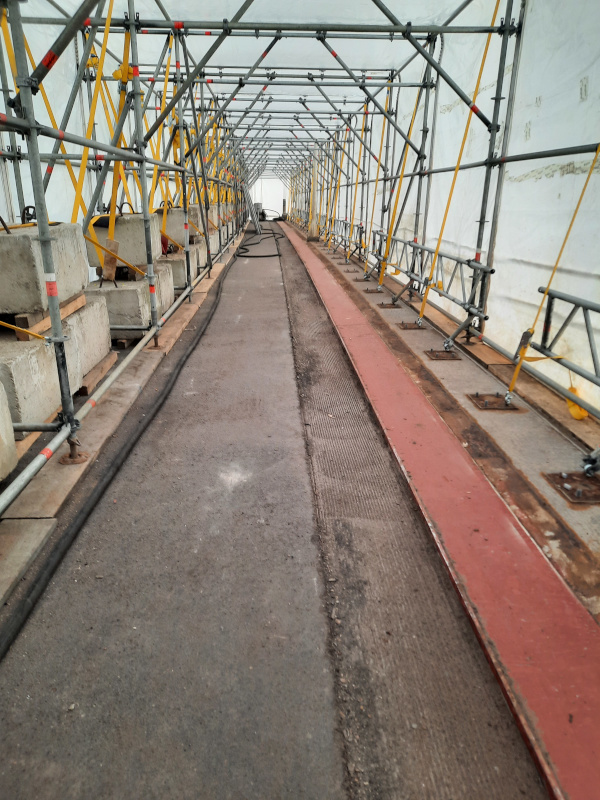 Über ein langegezogenes Gerüst liegt ein weiße Folie und bildet dadurch einen Tunnel. Auf dem Boden ist ein Stück Asphal abgefräst. 