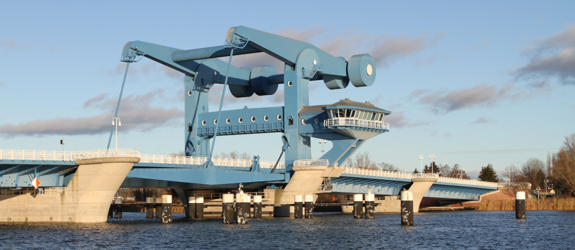 Eine blaue Stahl-Brücke erstreckt sich über einen Fluss, den Peenestrom. Das Wasser ist leicht bewegt, am Himmel sind ein paar Schleierwolken.