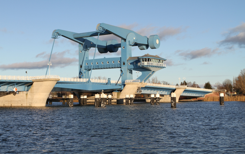 Eine blaue Stahl-Brücke erstreckt sich über einen Fluss, den Peenestrom. Das Wasser ist leicht bewegt, am Himmel sind ein paar Schleierwolken.