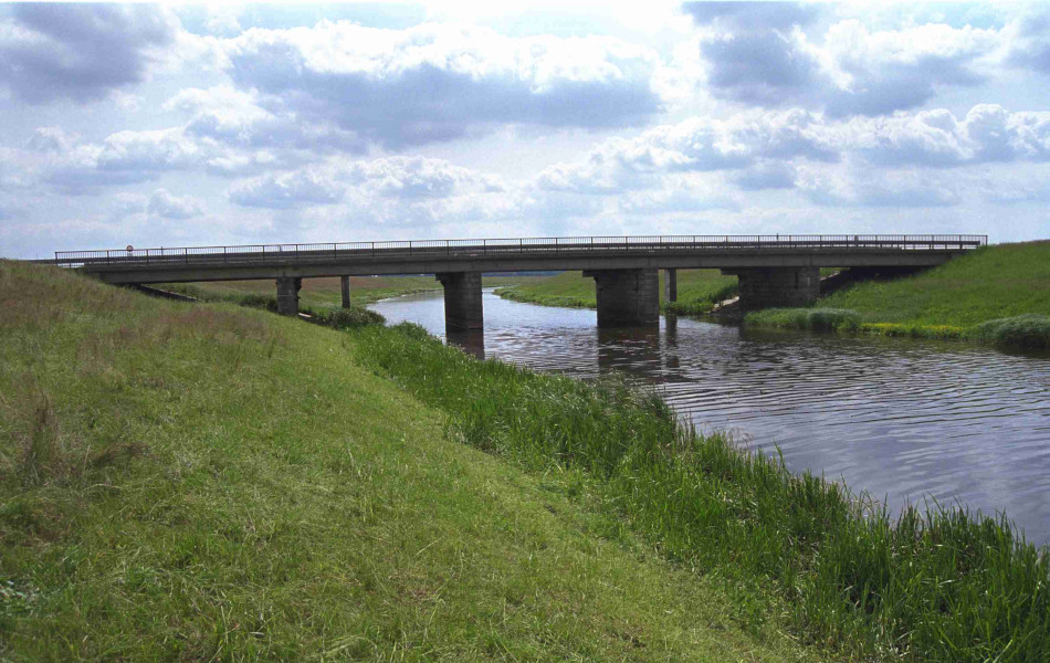 Eine Brücke mit Geländern führt über einen Fluss. In den Uferbereichen sind grüne Böschungen. 