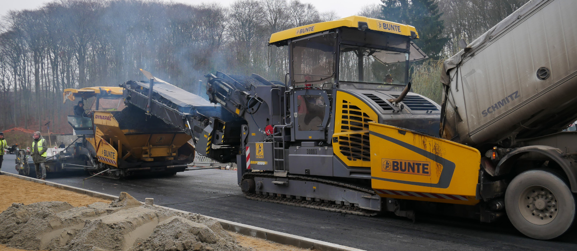 Baumaschinen bringen Asphalt auf eine Straße. Im Vordergrund sind eine Sandfläche und ein Sandhaufen.