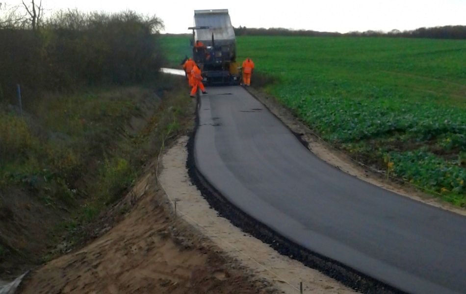 Ein frisch asphaltierter Weg führt an einem Feld entlang. Links neben dem Weg ist ein Gebüsch. Im Hintergrund ist eine Baumaschine mit Bauarbeitern zu sehen.