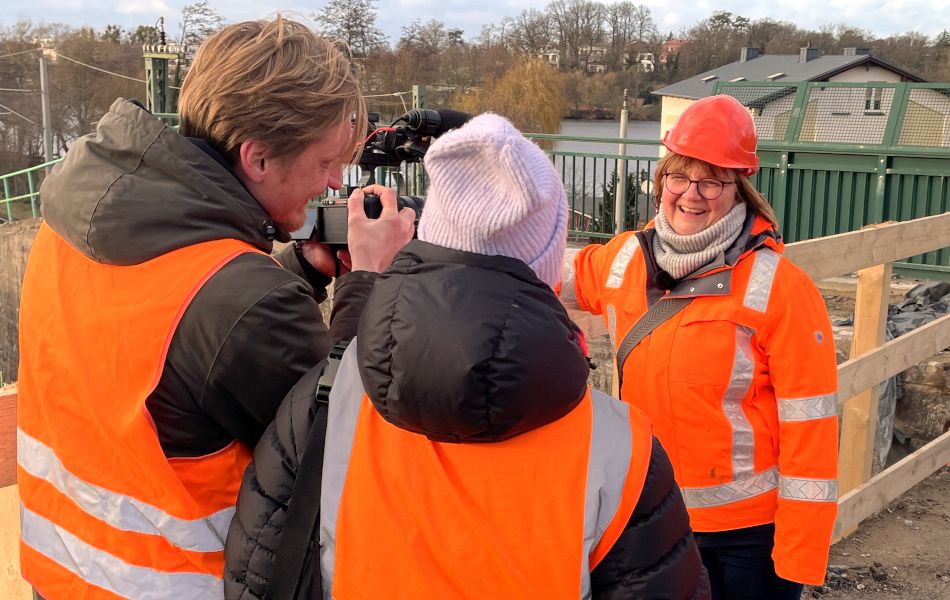 Ein Mann mit orangener Jacke und Bauhelm steht vor einer Kamera und gibt ein Interview. Im Hintergrund ist eine Baustelle.