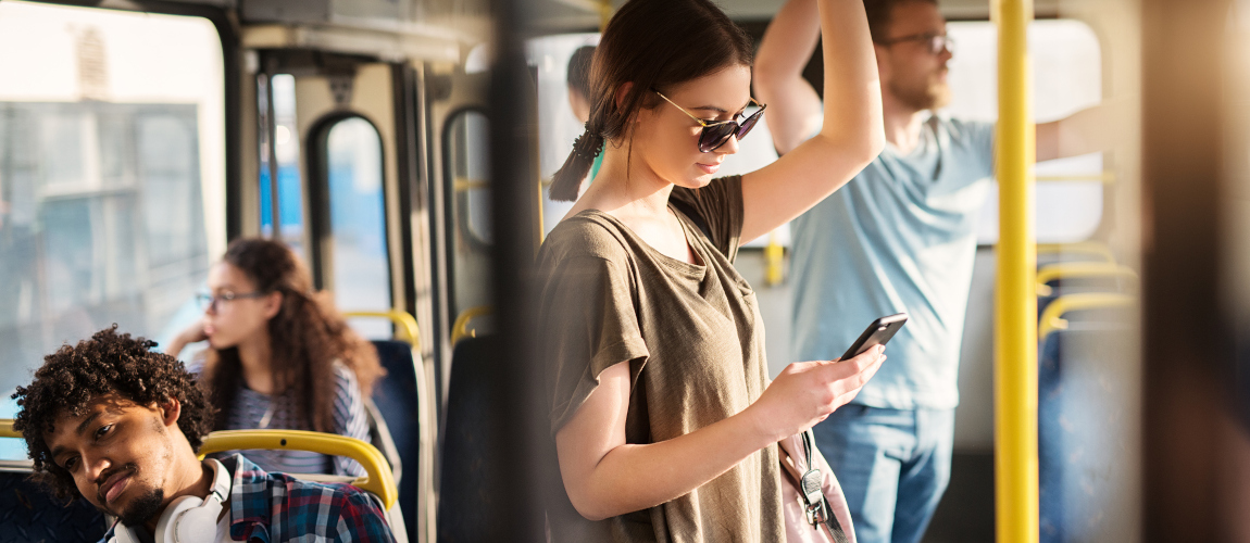 Eine junge Frau benutzt ihr Handy, während sie in einem Bus steht. Im Hintergrund sind weitere Fahrgäste.