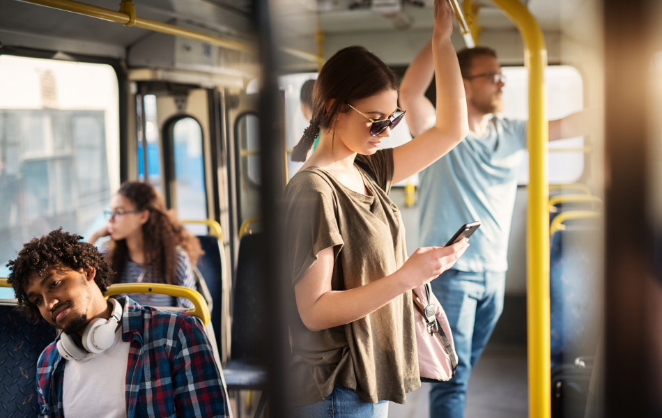 Eine junge Frau benutzt ihr Handy, während sie in einem Bus steht. Im Hintergrund sind weitere Fahrgäste.