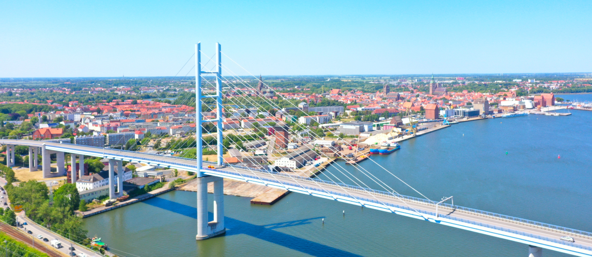 View from above of the city of Stralsund. In front of the city, a large road bridge with a huge pylon leads over a body of water, the Strelasund.
