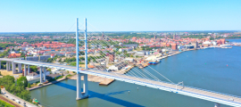 Blick von oben auf die Stadt Stralsund. Vor der Stadt führt eine große Straßenbrücke mit einem riesigen Pylonen über ein Gewässer, den Strelasund.