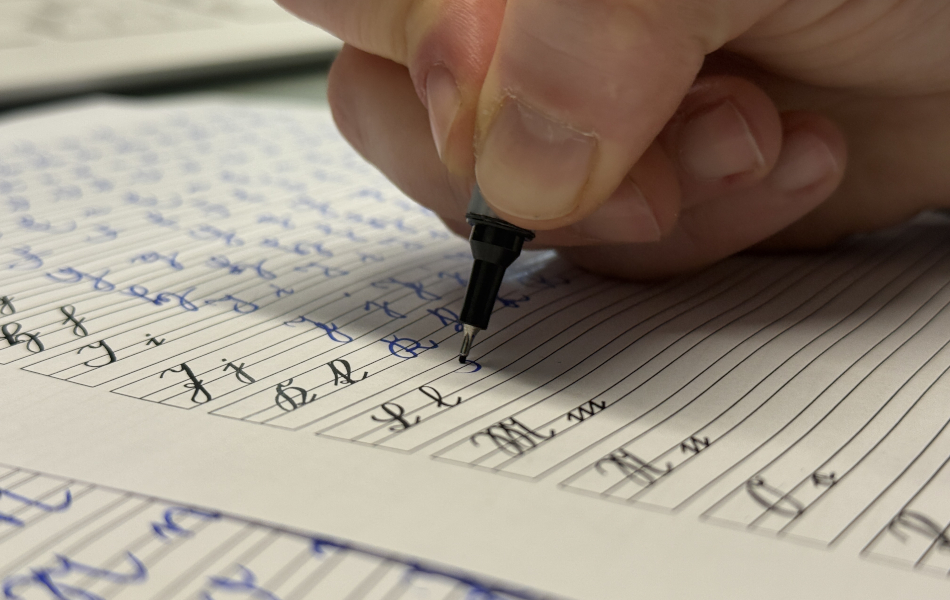 Close-up of a hand writing historical Kurrent letters with a pen on lined paper. Additional handwritten practice exercises are visible in the background.