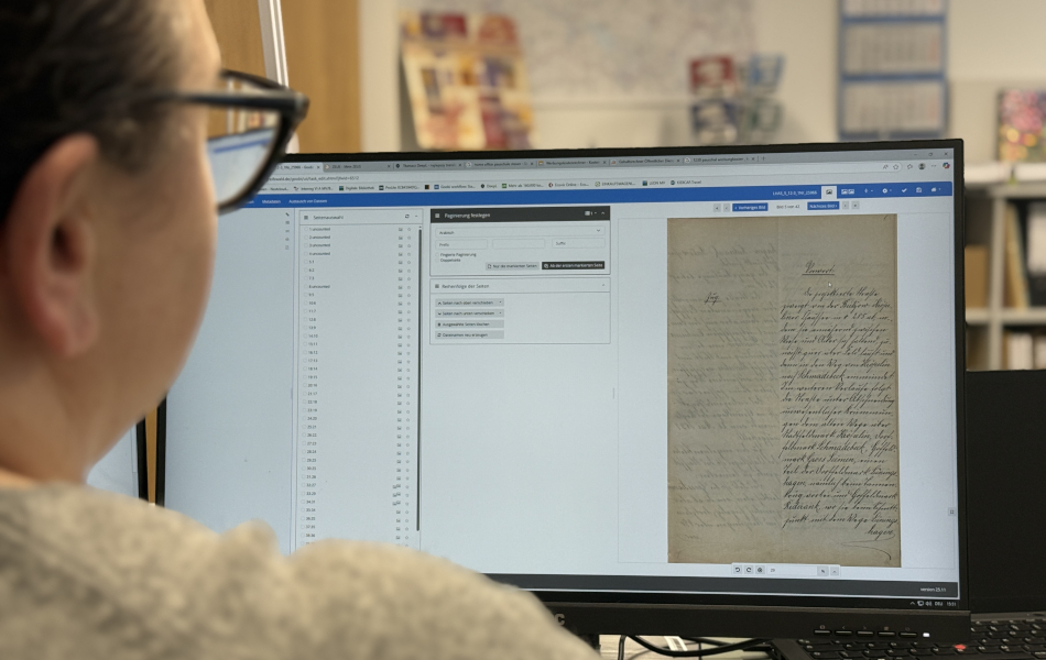 Eine Mitarbeiterin bearbeitet ein historisches handschriftliches Dokument am Computer. Auf dem Bildschirm ist eine gescannte Akte in einer Archivsoftware zu sehen.