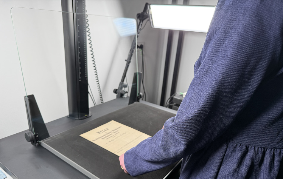 The image shows a person aligning a historical document on a flat reproduction surface. Above the document, a camera unit with lighting is mounted for contactless digitisation.