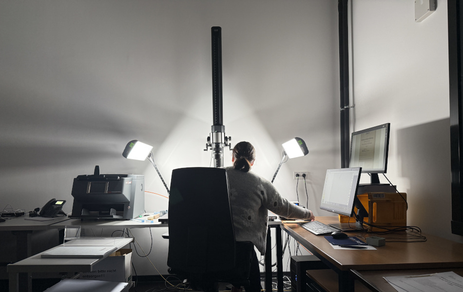 Eine Mitarbeiterin sitzt mit dem Rücken zur Kamera an einem Arbeitsplatz mit Scanner, zwei Leuchten und mehreren Bildschirmen.