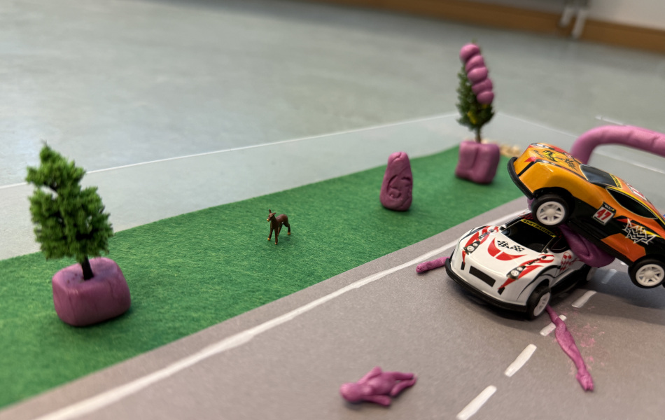 Selbstgebautes Straßenmodell mit Fahrbahn, Spielzeugautos, kleinen Bäumen und violetten Meilensteinen aus Knete am Straßenrand.