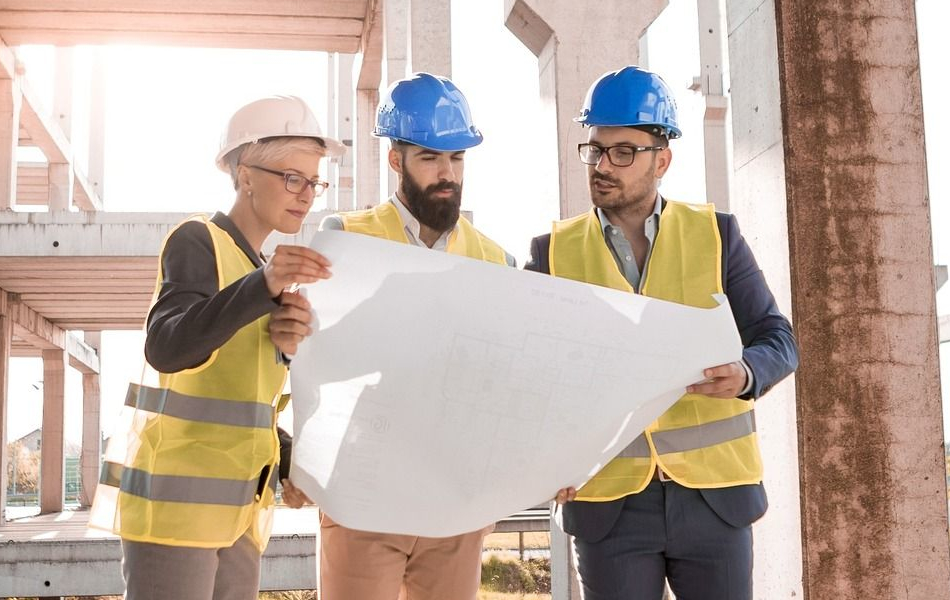 Zwei Männer und eine Frau mit Bauarbeiterhelm und gelber Signalweste halten gemeinsam einen Plan in den Händen und diskutieren darüber. Im Hintergrund ist ein Gebäude zu sehen, das sich in Bau befindet.
