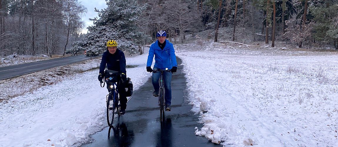 Zwei Männer fahren mit Fahrrädern auf dem neuen Radweg an der Bundesstraße. Auf den angrenzenden Grünflächen und Bäumen liegt Schnee.
