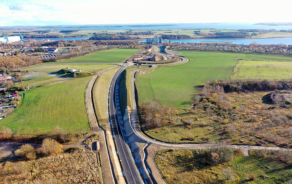 Luftaufnahme einer neuen Straßen­trasse mit Kurvenverlauf durch offene Felder bei Wolgast, im Hintergrund die Stadt und Wasserflächen.