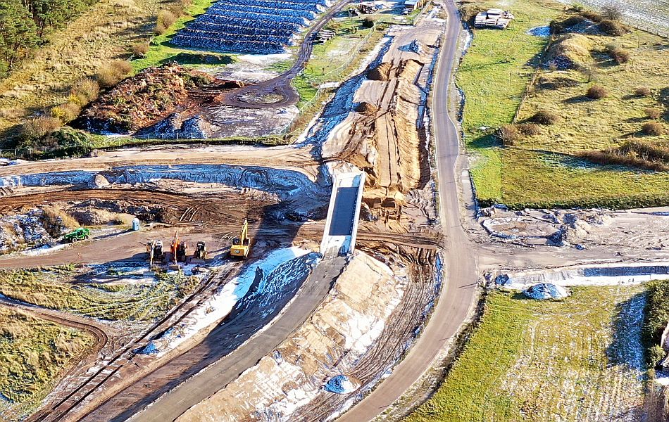Baustellenaufnahme mit Erdarbeiten und mehreren Baggern, mittig eine im Bau befindliche Brücke über eine Einschnitttrasse, umgeben von Wiesen und Lagerflächen.