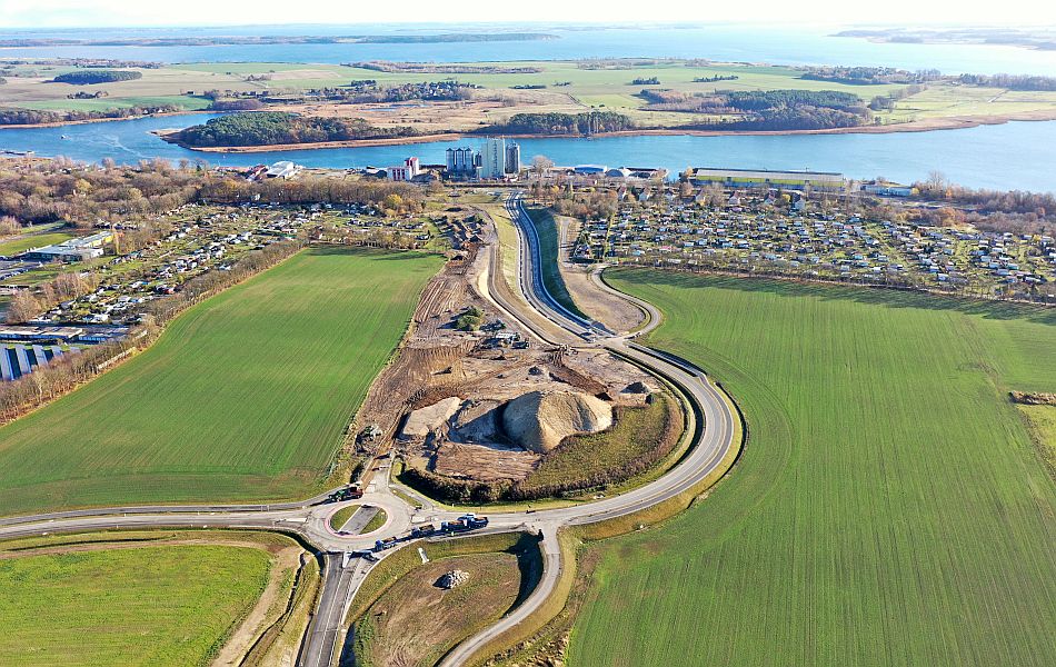 Luftbild eines Kreisverkehrs mit angeschlossenem Neubauabschnitt der Straße, dahinter grüne Felder, Baustellenflächen und das Peenestrom-Ufer.