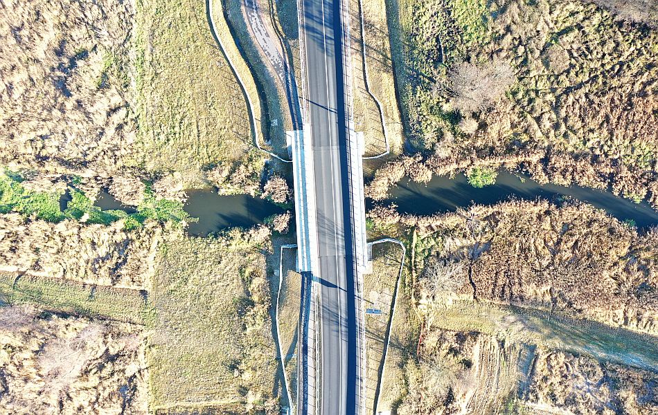 Draufsicht auf eine neue Straßenbrücke über einen schmalen Flusslauf, umgeben von Schilf und Grünflächen, klare geometrische Linienführung.
