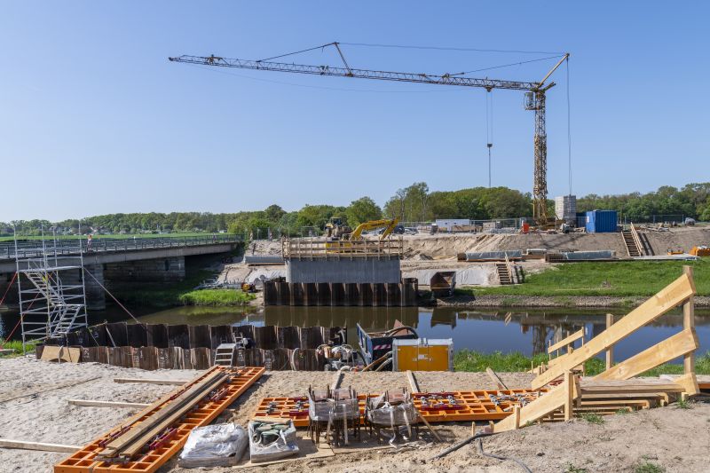 Auf einer Baustelle liegt am Ufer eines Flusses Baumaterial auf sandigem Boden, darunter unter anderem Schalplatten. Im Hintergrund stehen am Ufer auf der anderen Seite des Flusses der erste Betonpfeiler einer neuen Brücke und dahinter ein riesiger Kran.