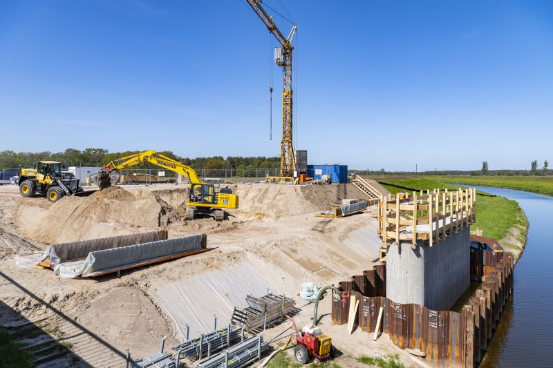 Auf einer Baustelle am Ufer eines Flusses schaufeln zwei Bagger an einem Sandberg. Rechts steht direkt am Wasser der erste Betonpfeiler einer neuen Brücke. Im Hintergrund ist ein riesiger Kran. © Matthäi-Gruppe Auf einer Baustelle am Ufer eines Flusses schaufeln zwei Bagger an einem Sandberg. Rechts steht direkt am Wasser der erste Betonpfeiler einer neuen Brücke. Im Hintergrund ist ein riesiger Kran.