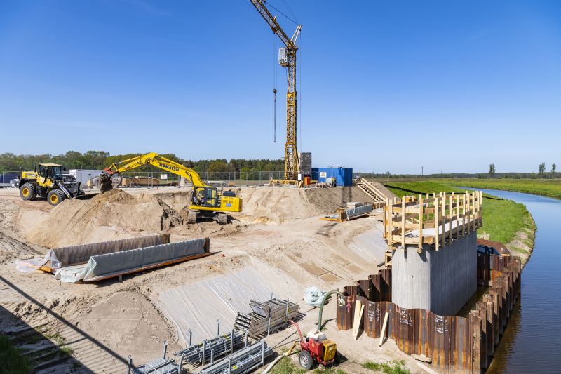 Auf einer Baustelle am Ufer eines Flusses schaufeln zwei Bagger an einem Sandberg. Rechts steht direkt am Wasser der erste Betonpfeiler einer neuen Brücke. Im Hintergrund ist ein riesiger Kran.