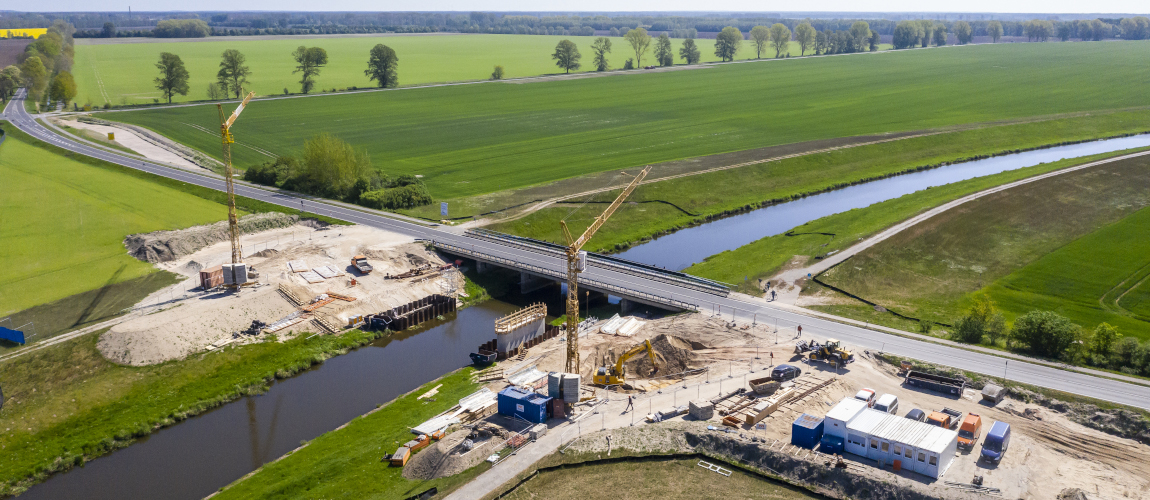 Blick aus luftiger Höhe auf eine Baustelle: Eine Straßenbrücke führt über einen Fluss. Direkt daneben entsteht eine neue Brücke, ein erster Betonpfeiler ist bereits fertig. An den Ufern des Flusses steht auf jeder Seite ein riesiger Kran.