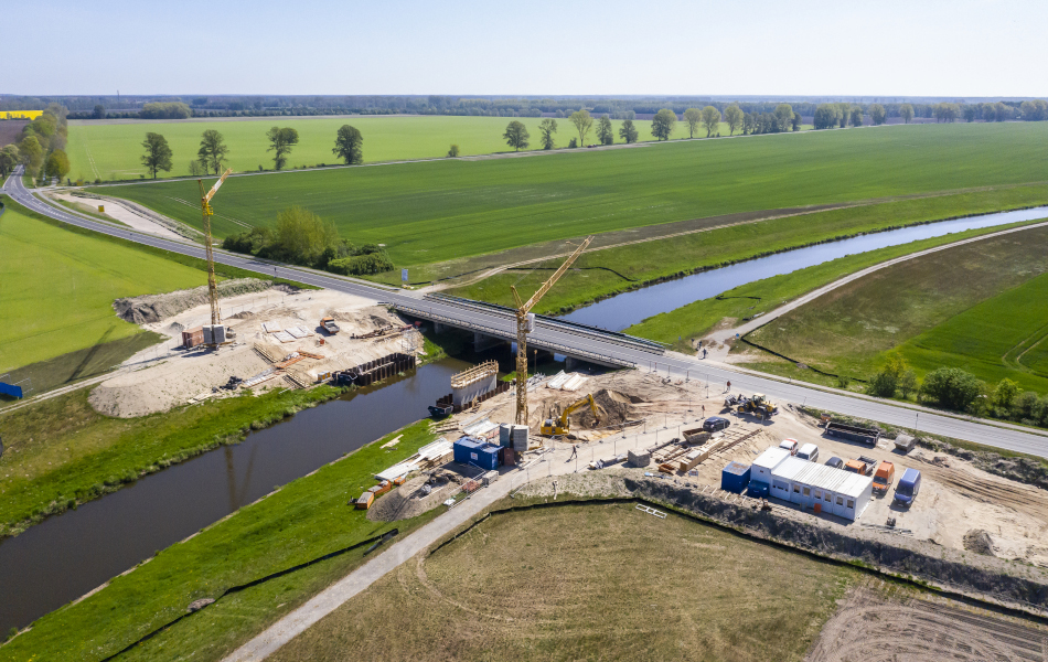 Blick aus luftiger Höhe auf eine Baustelle: Eine Straßenbrücke führt über einen Fluss. Direkt daneben entsteht eine neue Brücke, ein erster Betonpfeiler ist bereits fertig. An den Ufern des Flusses steht auf jeder Seite ein riesiger Kran.