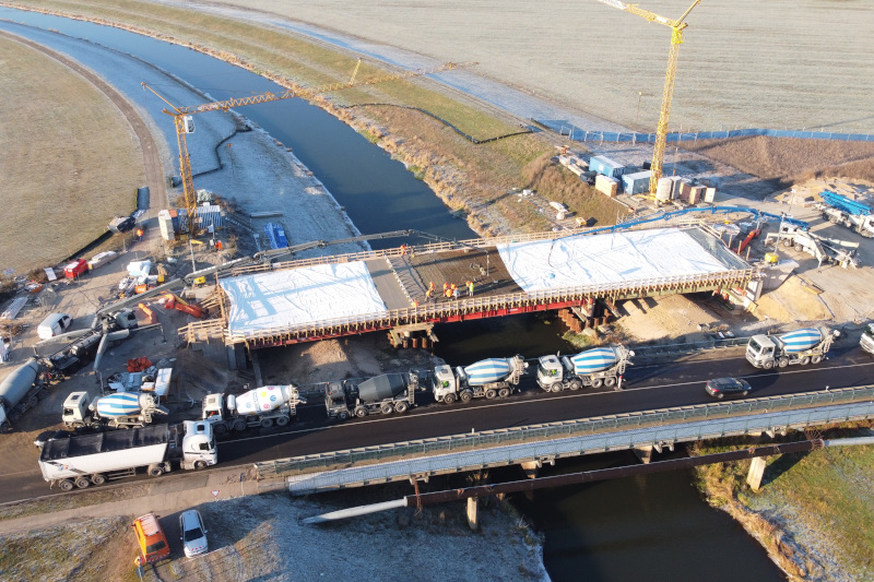 Blick auf eine Baustelle aus der Luft: Neben einer bestehenden Brücke, auf der mehrere Betonmisch-Lkw, wird eine neue Brücke errichtet. Mehrere Bauarbeiten stehen auf der neuen Brücke, die größtenteils von zwei riesigen weißen Planen zugedeckt wird. Neben der Brücke steht auf jeder Uferseite jeweils ein großer Kran.