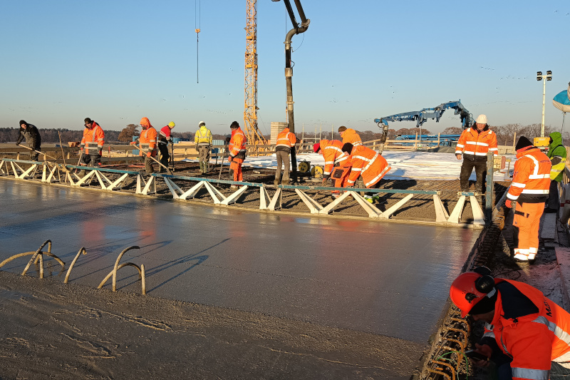 Mehrere Bauarbeiter stehen auf Eisenstäben und verteilen darin flüssigen Beton. Einen Teil der Betonage haben Sie bereits fertig und mit einem langen Holzstück glatt gezogen. Im Hintergrund steht ein Baukran.