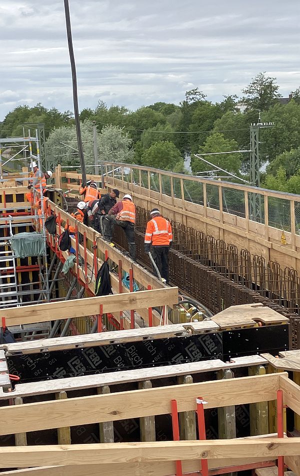 Mehrere Bauarbeiter sind auf einer hohen Baukonstruktion aus Holz und Stahl tätig. Im Hintergrund sind Bäume und Masten für die Fahrstromleitung einer Bahnstrecke.