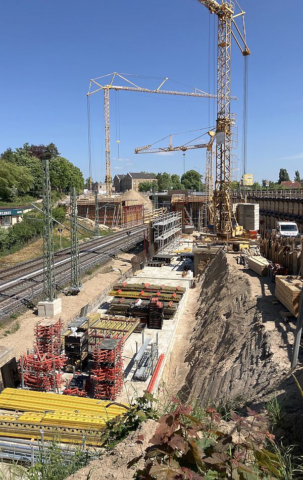 An einer Bahnstrecke befindet sich eine große Baustelle mit drei Kränen. Für den Neubau einer Brücke über die Bahngleise sind die ersten Baukonstruktionen aus Stahl und Holz entstanden. Am Rand der Gleise lagert reichlich Baumaterial. Im Hintergrund sind eine alte Brücke und Wohngebäude.