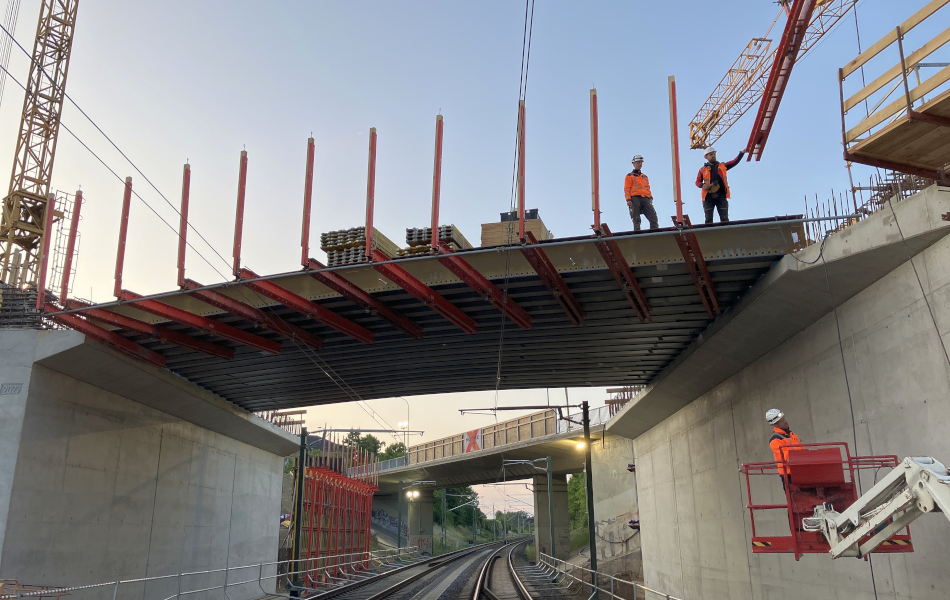Brückenbaustelle über einer Bahnstrecke: Zwei Bauarbeiter stehen auf einer teilweise montierten Stahlkonstruktion, ein dritter arbeitet von einer Hebebühne aus. Unter der Brücke verlaufen zwei Gleise mit Oberleitungen. Ein Kran hebt Bauteile an, auf der Brücke sind Baustoffe gelagert. © strassen-mv.de Brückenbaustelle über einer Bahnstrecke: Zwei Bauarbeiter stehen auf einer teilweise montierten Stahlkonstruktion, ein dritter arbeitet von einer Hebebühne aus. Unter der Brücke verlaufen zwei Gleise mit Oberleitungen. Ein Kran hebt Bauteile an, auf der Brücke sind Baustoffe gelagert.